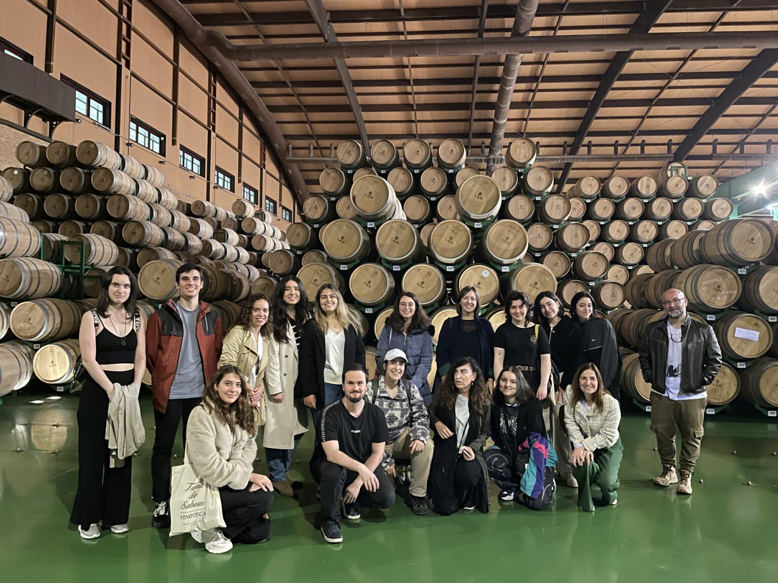 Grupa około siedemnastu osób stoi w winiarni przed stosami beczek z winem. Większość osób uśmiecha się do kamery. W tle widać duże drewniane beczki ułożone w kilku rzędach sięgających sufitu