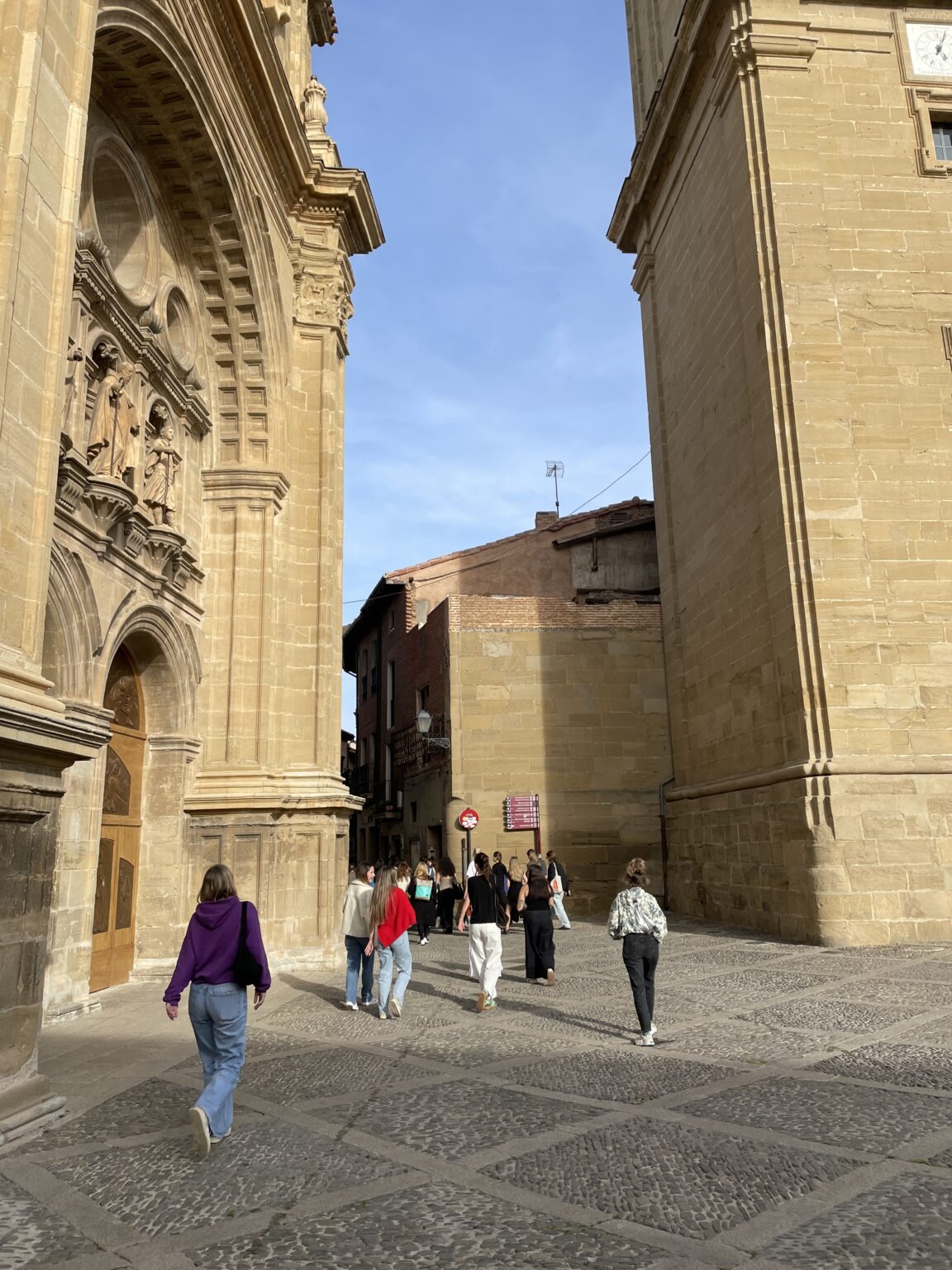 Grupa ludzi spaceruje po kamiennej ulicy pomiędzy dwoma dużymi, jasnobrązowymi budynkami o zabytkowej architekturze. Na pierwszym planie widać wysokie łuki i rzeźby zdobiące fasadę jednego z budynków. Niebieskie niebo i jasne światło słoneczne sugerują pogodny dzień.